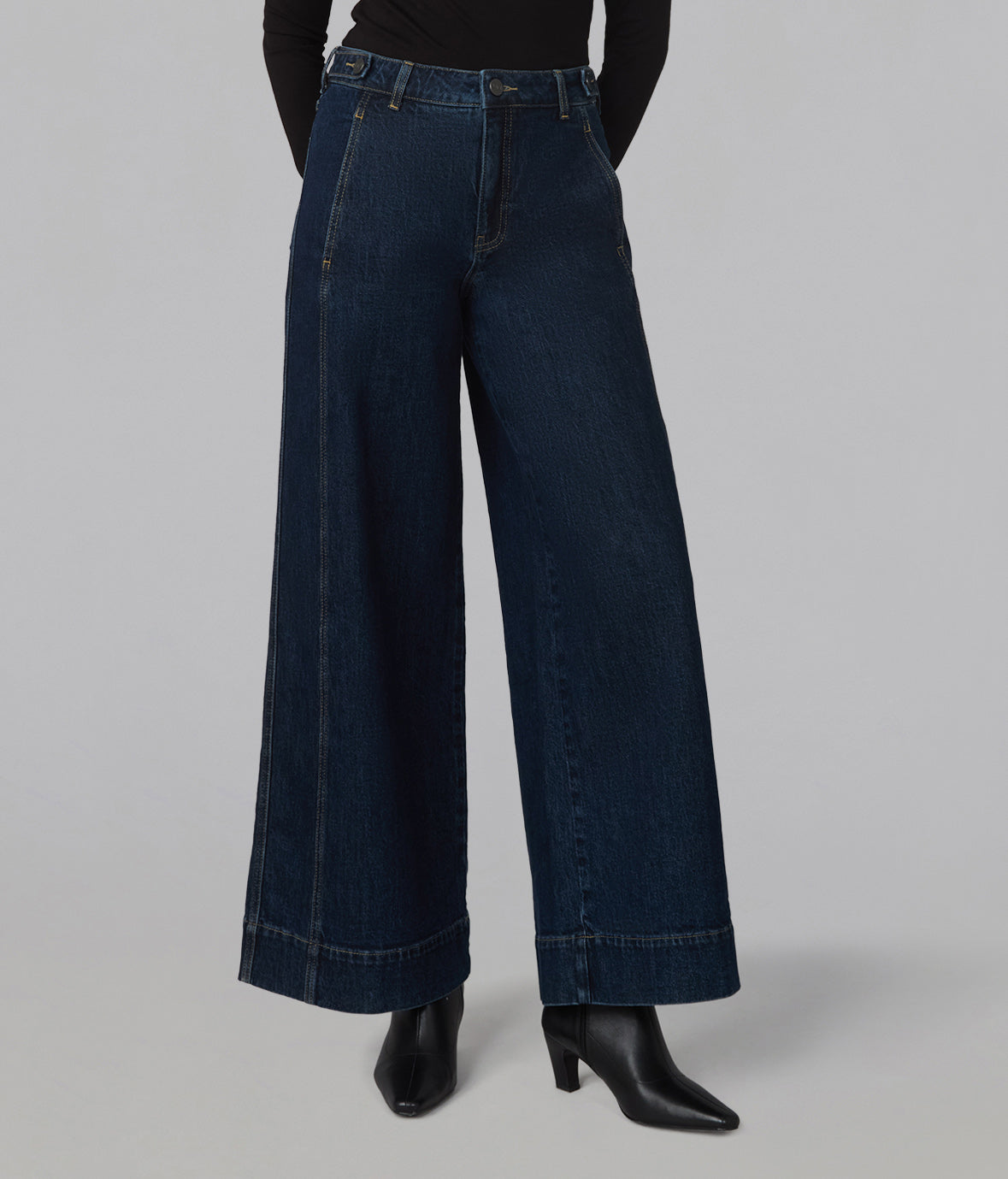 A model is shown from the waist down wearing dark blue wide-leg denim jeans, a black long-sleeve top, and black pointed-toe heeled boots against a solid gray studio background.